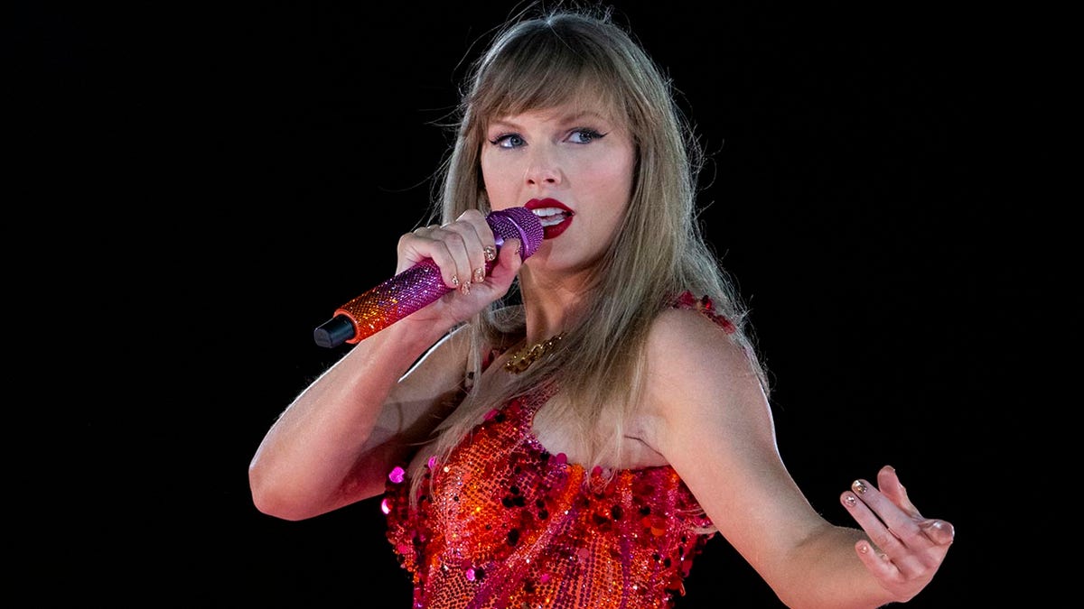 Taylor Swift canta frente a un deslumbrante micrófono naranja y morado y mira a su izquierda en el escenario mientras usa un mono rojo/naranja.