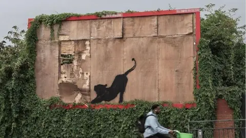 Banksy Una persona anda en bicicleta frente a una obra de graffiti de un gato grande tendido en un cartel de madera.